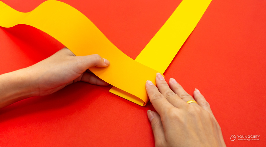 พับกระดาษสีส้มจากด้านขวาไปทางด้านซ้ายให้ทับกระดาษสีเหลือง