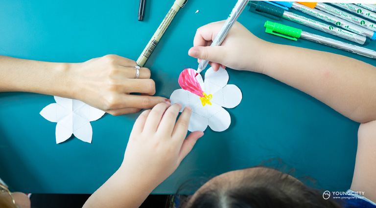 เด็กและผู้ปกครองช่วยกันคลี่กระดาษออกมาแล้วระบายสีดอกไม้ให้สวยงาม