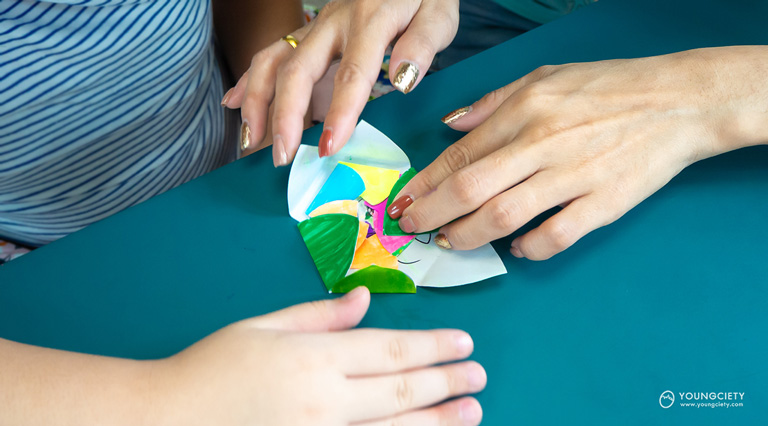 เด็กและผู้ปกครองนำดอกไม้กระดาษที่พับเสร็จแล้ว นำมาซ้อนกันบนกระดาษใบไม้กระดาษและพับใบไม้ทับอีกชั้น
